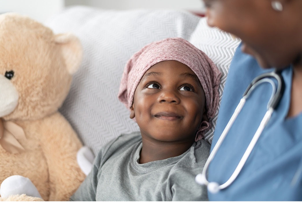 child with cancer in bed with stuffed animal and nurse sitting nearby