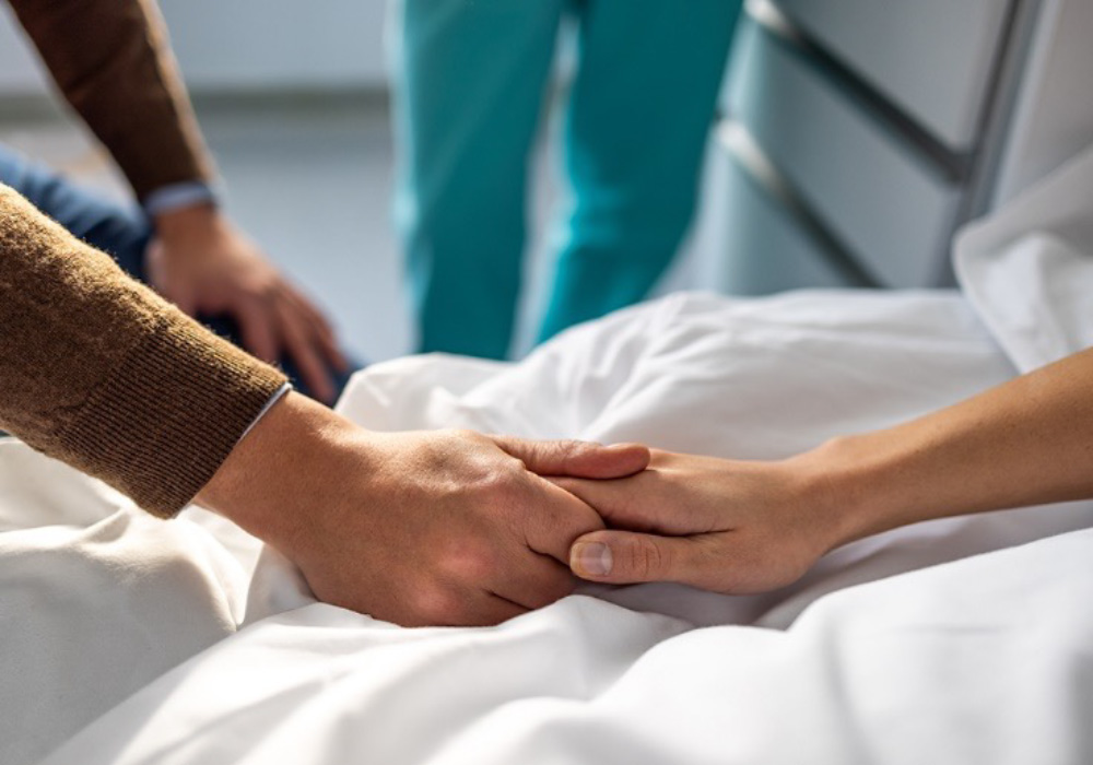 person with brown clothes holding patient's hand in support on hospital bed 