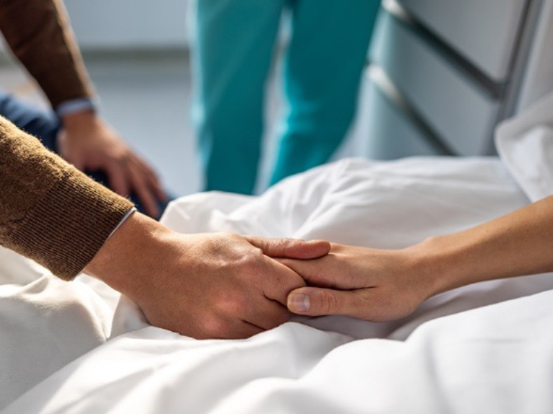person with brown clothes holding patient's hand in support on hospital bed 
