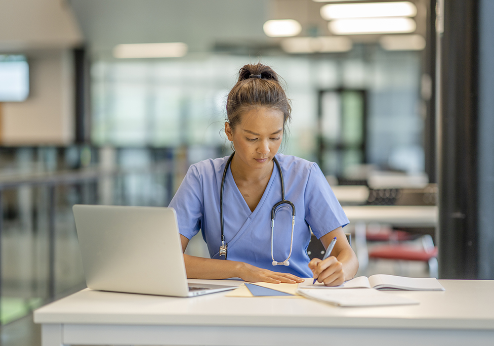 nurse in scrubs sitting at a desk with a computer and taking notes