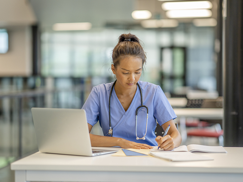 nurse in scrubs sitting at a desk with a computer and taking notes