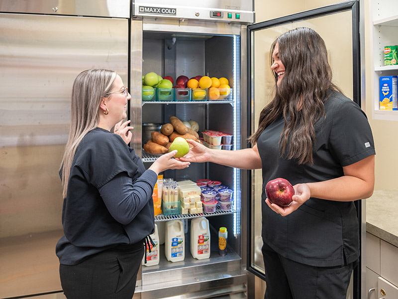 Nurse handing fruit to another nurse at refrigerator